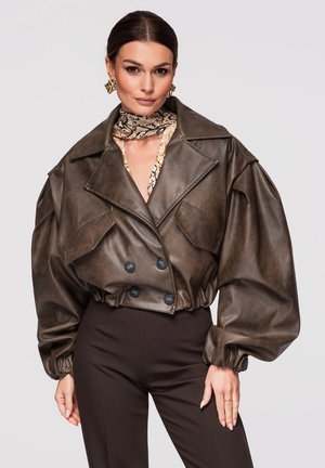 Žena nosící velkou tmavě hnědou koženou bundu s nafouklými rukávy, šátek s hadím vzorem, zlaté náušnice a přiléhavé tmavé kalhoty.