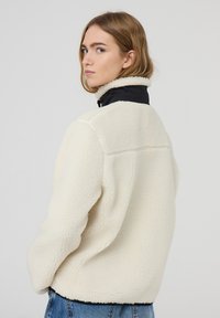 Giacca in pile crema con colletto alto e dettagli neri, caratterizzata da una superficie testurizzata e una vestibilità comoda. Jeans di denim visibili sotto.