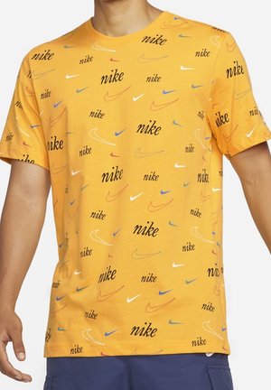 Gul kortærmet t-shirt med gentagne multicolor Nike-logoer og swoosh-mønstre båret af en person mod en ensfarvet baggrund.