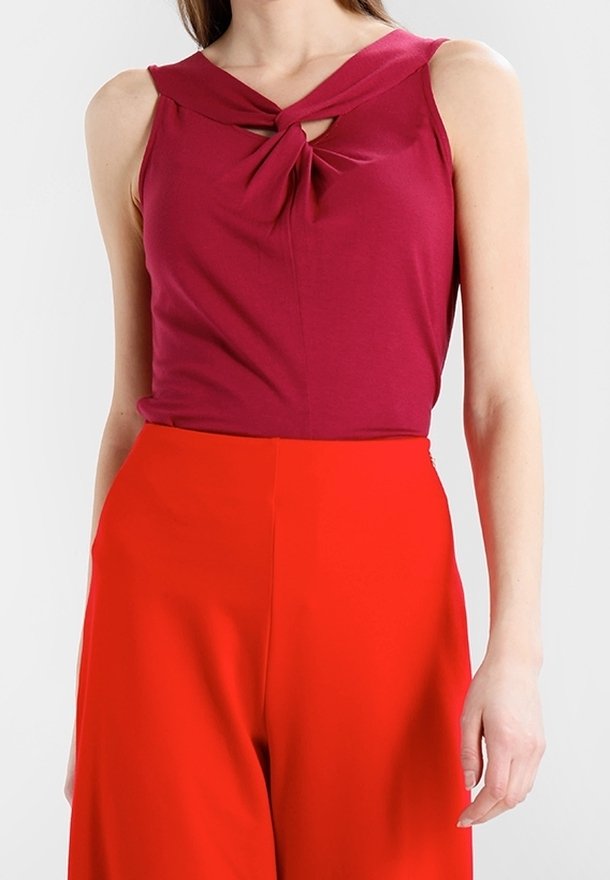 Žena v bezrukávovém tmavě růžovém topu s krouceným výstřihem a vysoko sedícími jasně červenými širokými kalhotami na bílém pozadí.