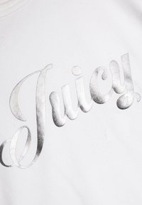 Biely textil s lesklým strieborným logom "Juicy" v zdobenom písmene. Hladká textúra s podrobnými krivkami a akcentmi v písaní.