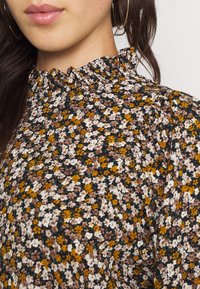 Blusa floreale con collo volant, sfondo nero, piccoli fiori bianchi, gialli e marroni, con un tessuto strutturato e maniche a sbuffo.