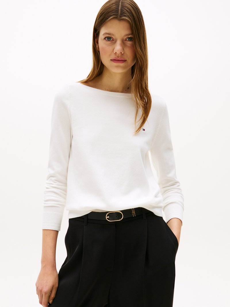 Femme portant un haut à manches longues blanc et un pantalon noir avec une ceinture noire, se tenant avec une main dans sa poche devant un fond uni.