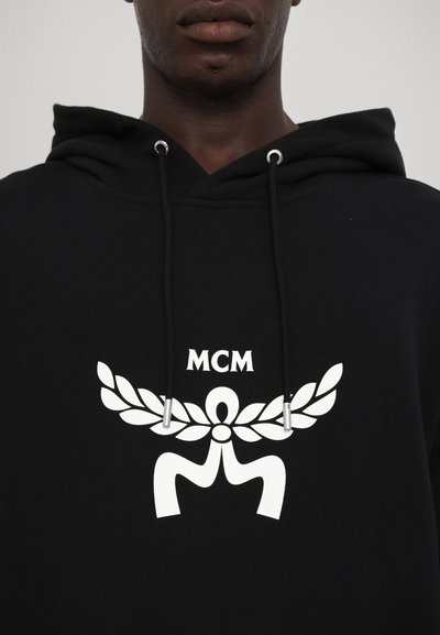 Hoodie noir avec une poche kangourou, présentant un grand logo graphique blanc avec un design de laurier. Des cordons avec embouts métalliques complètent le look.