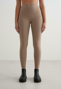Beige augstās jostas leggings, izgatavoti no elastīga materiāla, ar gludu tekstūru un logotipu jostasvietā. Valkāti ar melniem potīšu zābakiem.