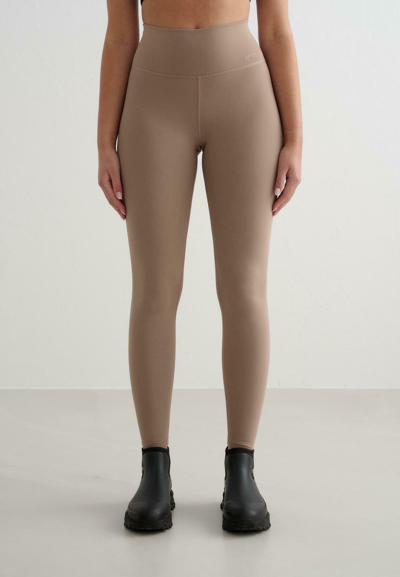 Beige augstās jostas leggings, izgatavoti no elastīga materiāla, ar gludu tekstūru un logotipu jostasvietā. Valkāti ar melniem potīšu zābakiem.