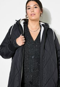 Černá prošívaná puffer bunda s kapucí, s bílou umělou kožešinou na vnitřní straně a kontrastním svetrem pod ní, zdobená černými knoflíky.