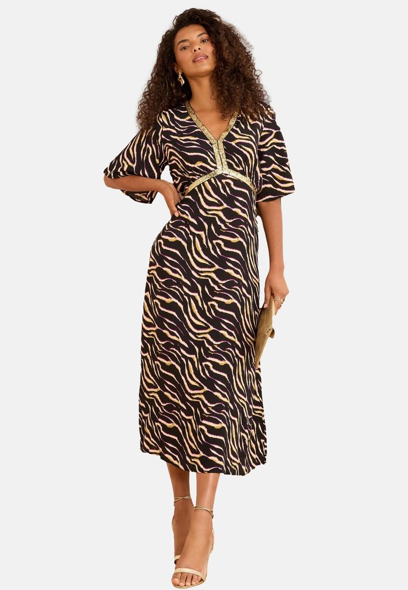 Kvinde iført en sort og beige midi kjole med tigerstriber og guldkant, holder en clutch og har nøgen ankelsandaler på.