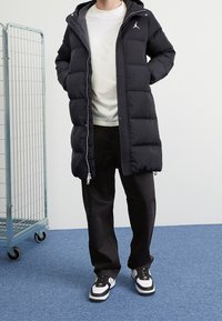 Zwarte pufferjas met capuchon, ritsluiting en horizontale stiksels. G gedragen over een wit shirt en gecombineerd met donkere broeken en zwarte sneakers.