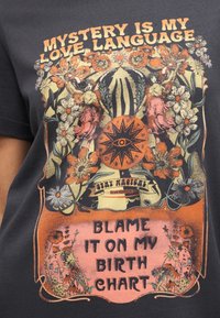 Το κείμενο «Το μυστήριο είναι η γλώσσα της αγάπης μου» και «Φταίει το αστρολογικό μου χάρτη» περικυκλώνει ένα πανόραμικο μάτι, λουλούδια και δύο γυναίκες που χορεύουν σε σκοτεινό πουκάμισο.