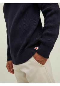 Jersey de punto de color azul marino con canalé, de mangas largas y un pequeño detalle de logo rojo y blanco en el dobladillo, combinado con pantalones crema.