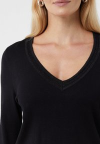 Schwarzer Pullover mit tiefem V-Ausschnitt und strukturiertem, metallic schimmerndem Saum. Der Stoff ist glatt und die taillierte Passform betont den Ausschnitt.