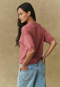 Rožnat pulover s kratkimi rokavi in ovratnikom z ohlapnim krojem, kombiniran z svetlo modrimi jeans hlačami širokih nogavic z vidnimi zadnjimi žepi.