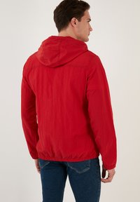 Jachetă roșie cu glugă, cu o textură ușoară și crinkled. Are elastic la manșete și la tiv, și un design simplu. Fără modele sau accente vizibile.