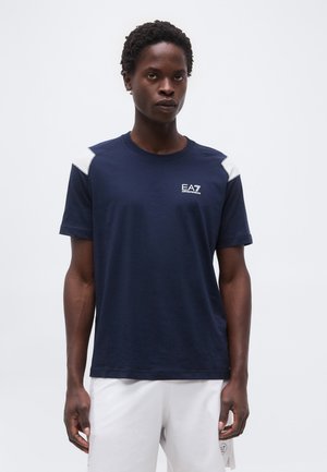 Férfi sötétkék EA7 Emporio Armani pólóban, fehér vállrészletekkel és fehér rövidnadrágban, egyszínű háttér előtt áll.