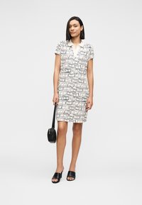 Φόρεμα με κοντά μανίκια και γιακά σε κρεμ χρώμα με γεωμετρικό μαύρο σχέδιο. Το μοντέλο κρατάει μια μικρή μαύρη τσάντα και φοράει μαύρα σανδάλια slides.