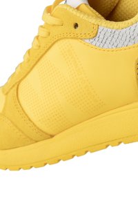 Scarpa sportiva gialla con una parte superiore in sintetico liscio decorata con dettagli testurizzati, colletto imbottito e suola in gomma scanalata.