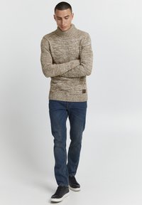 Strikket genser i beige med høy ripplet krage, teksturert mønster, akkompagnert av mørkeblå jeans og sorte sneakers med hvite såler.