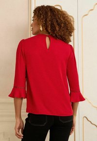 Blusa rossa con maniche a tre quarti, polsini svasati e dettaglio a chiave sulla schiena. Tessuto liscio con sottili decorazioni vicino ai polsini.
