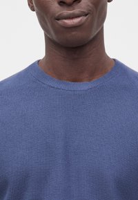 Gros plan d'une personne portant un pull tricoté bleu texturé, montrant le bas du visage, le cou et le haut de la poitrine sur un fond uni.