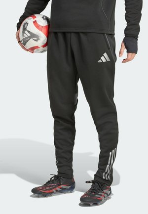 Personne en tenue de sport noire tenant un ballon de football rouge et blanc, portant un pantalon de survêtement Adidas noir et des crampons noirs.