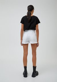Femme aux longs cheveux foncés attachés en queue de cheval, portant un t-shirt noir, un short blanc à ourlet roulé et des bottines noires épaisses, debout devant un fond uni.