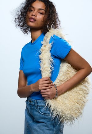 Mlada ženska z kodrastimi lasmi, oblečena v svetlo modro majico in jeans krilo, ki drži veliko puhasto ramo v krem barvi.