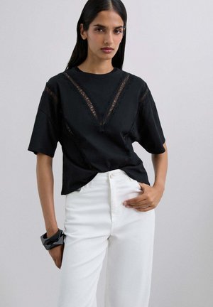 Camiseta negra de manga corta, con detalles de encaje en forma de V en la parte delantera. Combinada con unos vaqueros blancos de cintura alta y una pulsera negra.