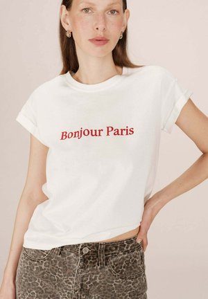 T-shirt bianco in cotone con maniche arrotolate, caratterizzato da testo rosso "Bonjour Paris". Indossato con jeans a stampa leopardata marrone e nera.