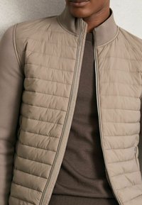 Beige quiltet bomberjakke med lynlås foran, stående krave og kontrastfarvede glatte ærmer, båret over en mørkebrun sweater.