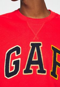 Primo piano di una persona che indossa una felpa rossa con grandi lettere in rilievo "GAP" in blu navale, crema e giallo cucite sul petto.