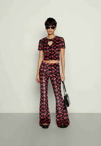 Top cropped preto e calças largas com um padrão de cerejas, apresentando um decote em forma de coração e textura brilhante. Modelo segura uma pequena bolsa preta.