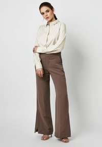 Cream and beige striped shirt with long sleeves, paired with high-waisted brown trousers featuring a relaxed fit and side slits.