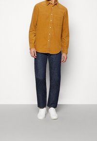 Senapsgul kordflorsskjorta med knapp-ned krage och bröstficka, i kombination med mörkblå jeans och vita sneakers. Slät textur och avslappnad passform.