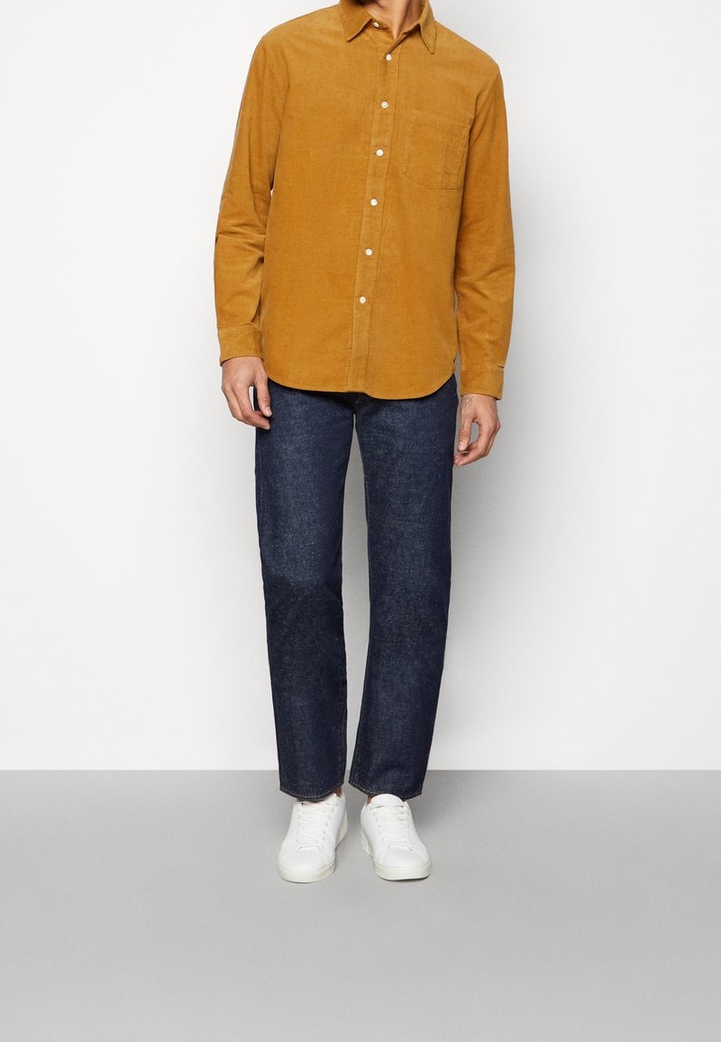 Senapsgul kordflorsskjorta med knapp-ned krage och bröstficka, i kombination med mörkblå jeans och vita sneakers. Slät textur och avslappnad passform.