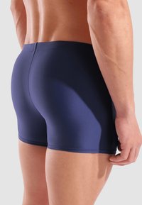 Marineblaue Badeshorts mit glatter Textur, einer figurschmeichelnden Passform und einer Länge bis zur Mitte des Oberschenkels. Sie verfügen über einen nahtlosen Bund und keine zusätzlichen Muster.