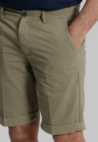 Pantaloni shorts verde oliva chiaro in cotone, con tasche laterali, chiusura con bottone e orlo arrotolato. Texture liscia con cuciture minime.