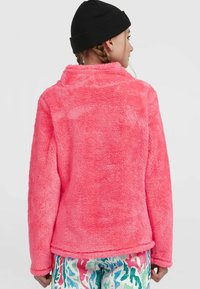 Fuzzy, lys pink fleece-jakke med høj krave, lange ærmer og en blød tekstur. Kombineret med mønstrede, farverige bukser.