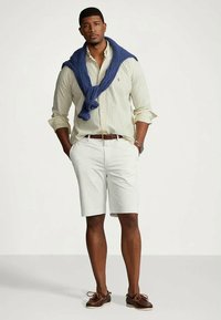 Άνδρας φορεμένος με πουκάμισο ριγέ, με τα μανίκια σηκωμένα, λευκά σορτς, καφέ ζώνη και παπούτσια, με ένα πουλόβερ ριγμένο στους ώμους, έχοντας τα χέρια του στις τσέπες.