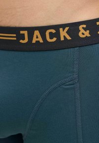 Férfi, testhezálló boxer alsó, téglalap formájú, türkiz színben, fekete, rugalmas derékpánttal, amelyen a "JACK &" felirat arany színben látható. Sima anyag, megerősített varrással.