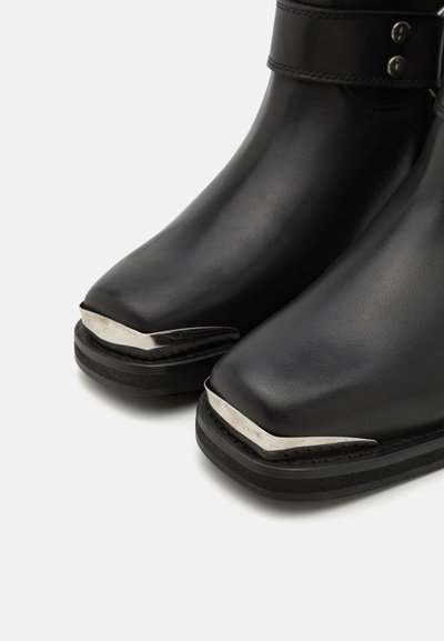 Botas de cuero negras con punta cuadrada y acentos metálicos en la parte frontal. Tienen una suela gruesa y una correa con hebilla alrededor del tobillo.