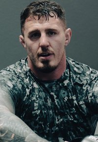 Atleta vistiendo una camiseta gris de camuflaje ajustada y de mangas cortas, con tatuajes visibles en los brazos y el cabello mojado, sentado contra un fondo gris.