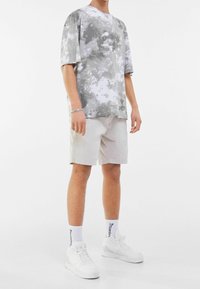 T-shirt tie-dye gris et blanc à manches courtes, associé à un short beige et des baskets blanches. La tenue a une coupe décontractée et un style casual.