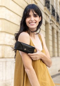 Étui à lunettes en cuir noir avec un design repliable, fermeture à pression en argent, tenu contre un bras nu. L'arrière-plan présente des éléments architecturaux beiges.