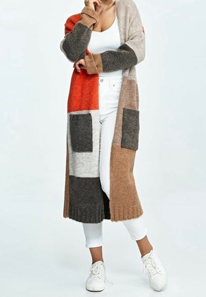 Mujer con cárdigan largo de bloques de color en naranja, beige, gris y marrón sobre camiseta blanca, pantalones blancos y zapatillas blancas.