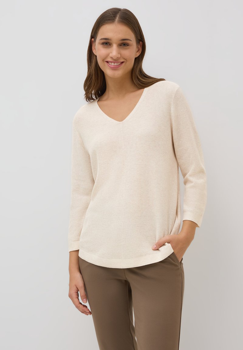 Krämfärgad stickad tröja med V-hals och trekvartslånga ärmar, med en mjuk textur och en avslappnad passform. Matchas med bruna byxor.