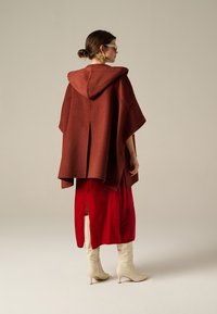 Poncho in lana color ruggine con cappuccio, caratterizzato da un design senza cuciture, indossato sopra un vestito di raso rosso, abbinato a stivali alti fino al ginocchio di colore off-white.
