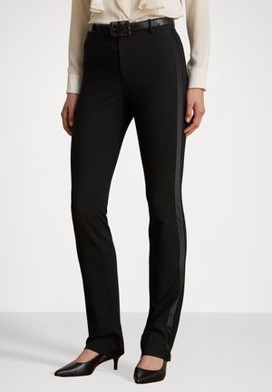 Schwarze taillierte Hose mit schmaler Passform, ausgestattet mit einem glänzenden Seitenstreifen und einer glatten Textur. Kombiniert mit einer weißen Bluse und schwarzen Absätzen.
