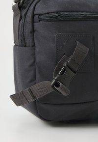 Borsa in tessuto nero con superficie texturizzata, dotata di chiusura con zip, tracolla regolabile e fibbia in plastica per il fissaggio.
