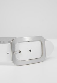 Hvid læderbælte med en rektangulær metallic sølvspænde med en glat overflade og en nålelukning. Justerbar med flere huller.